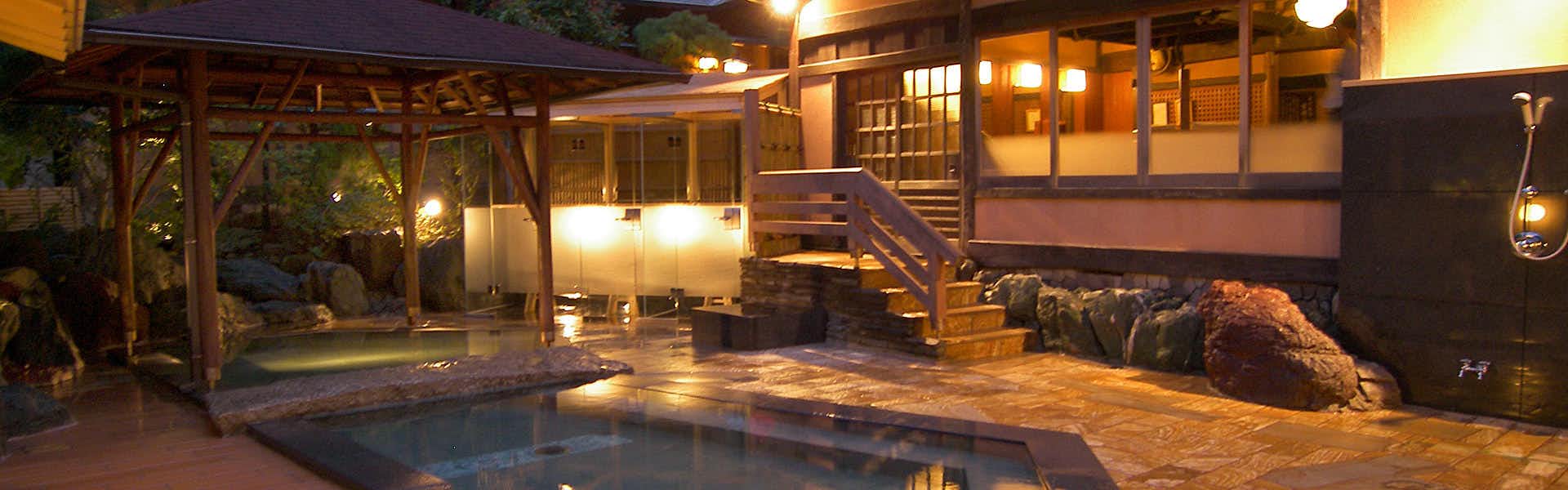Atami Sekitei exterior structure with wooden railings and platforms, illuminated lantern-style fixtures mounted on wooden framework, wooden deck, reflective water surface in foreground, wooden beams and structural elements