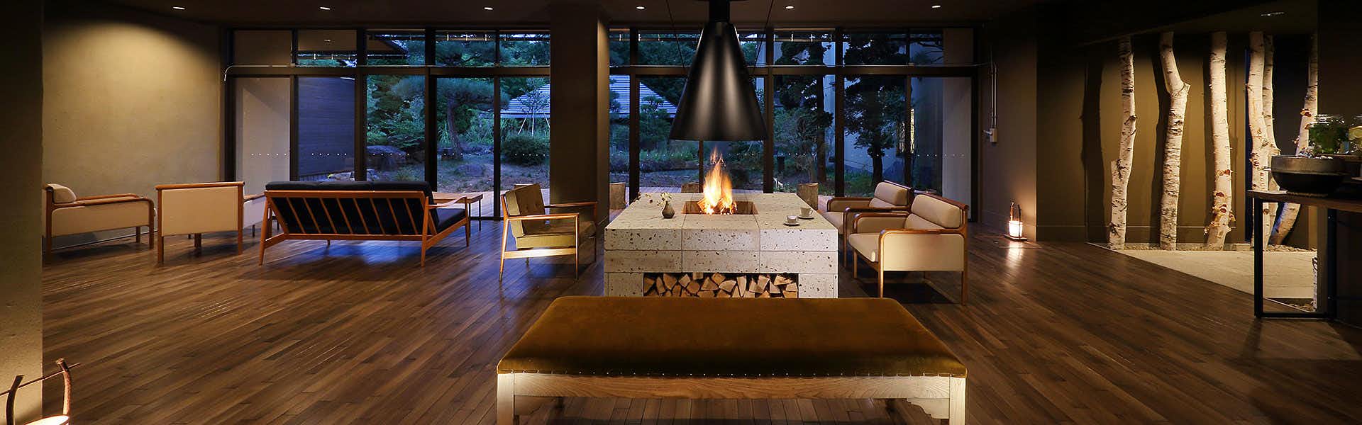 Oyado Mori no Ne Interior with dark wooden flooring, large windows showing forest views, white central fireplace, and seating areas.