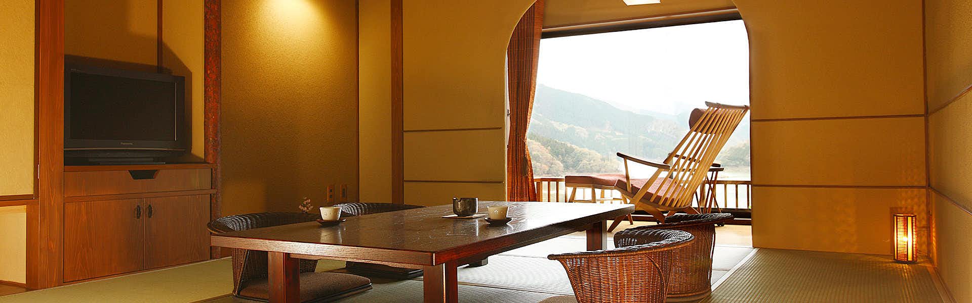 Akane Saian Sansui Interior with wooden furnishings and warm ochre walls, opening to a wooden deck overlooking a forested valley landscape.