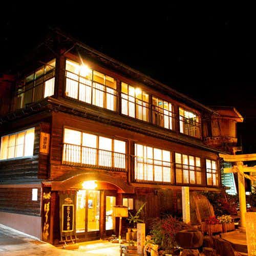 Horieya Ryokan Nighttime exterior illuminated in golden tones, two-story wooden building, white-framed glowing windows, yellow torii gate on right side.