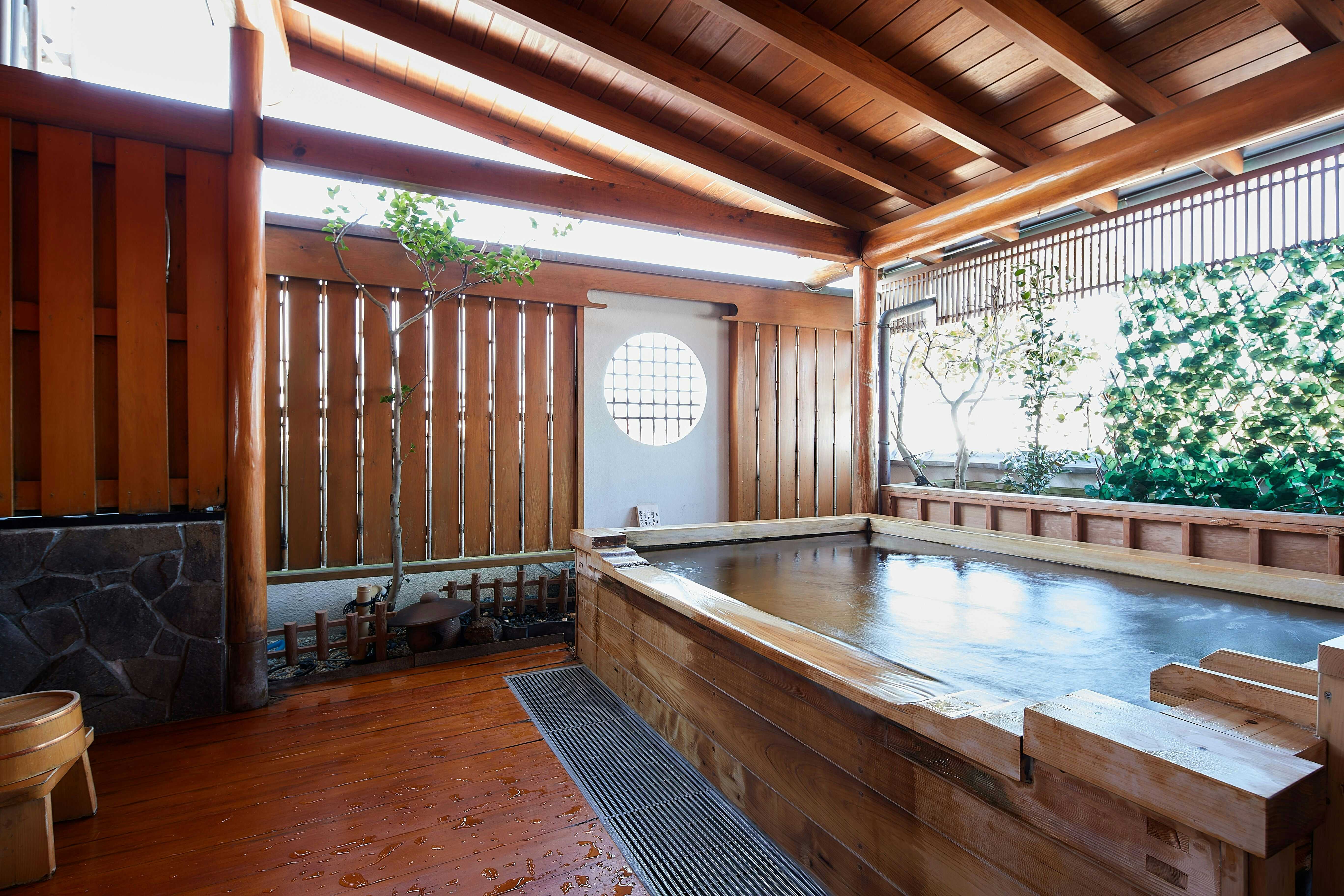 Aji no Yado Hanatsubaki Interior space with dark wooden sliding door panels with vertical slats, exposed wood beam ceiling, wooden bench seating, and window openings with climbing green plants outside.