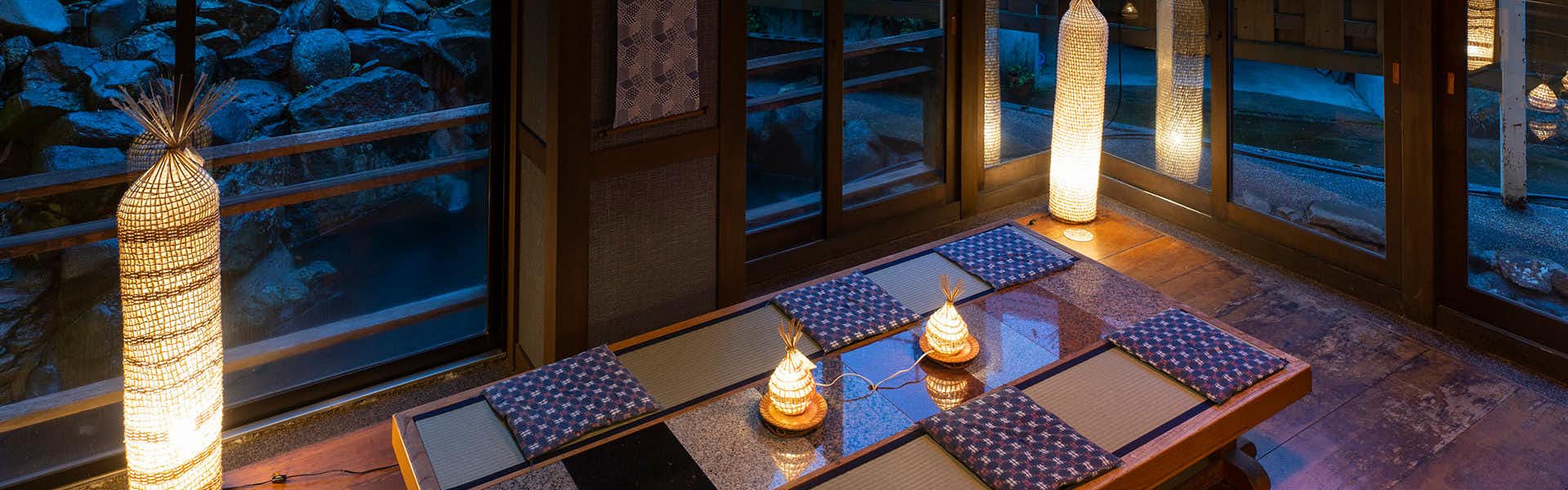 Nozawa Onsen Kawahichi-ya Ryokan Interior with woven pendant lamp, warm table lanterns, blue patterned cushions, and large windows with dark sky visible.