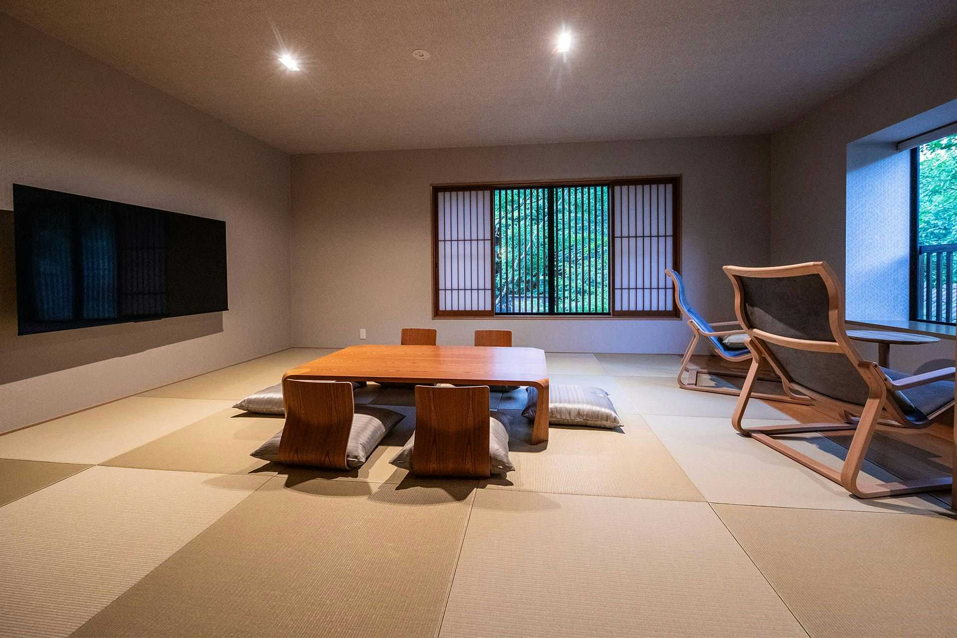 Yufuin Onsen Onyado Ichizen Tatami sitting room with low wooden table, floor cushions, two reclining chairs, wall-mounted television, and windows facing green trees