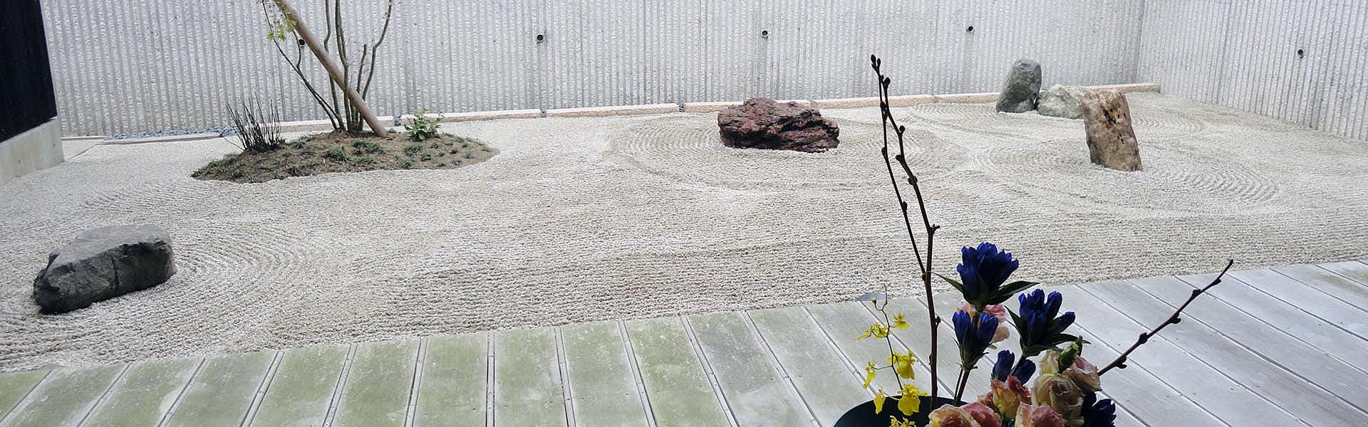 Himakajima Island Villa Yuzuki Gravel courtyard with scattered dark stones, green plants with dark foliage, corrugated boundary walls.