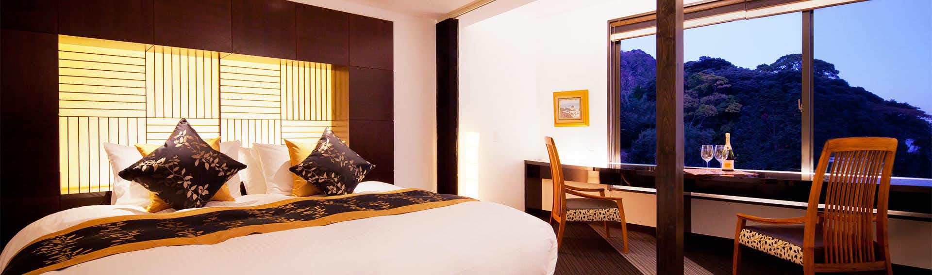 Isaribi Inn with All Rooms Featuring Open-Air Hot Spring Baths Bedroom with white linens, dark patterned cushions, and expansive windows framing mountain landscape at night.