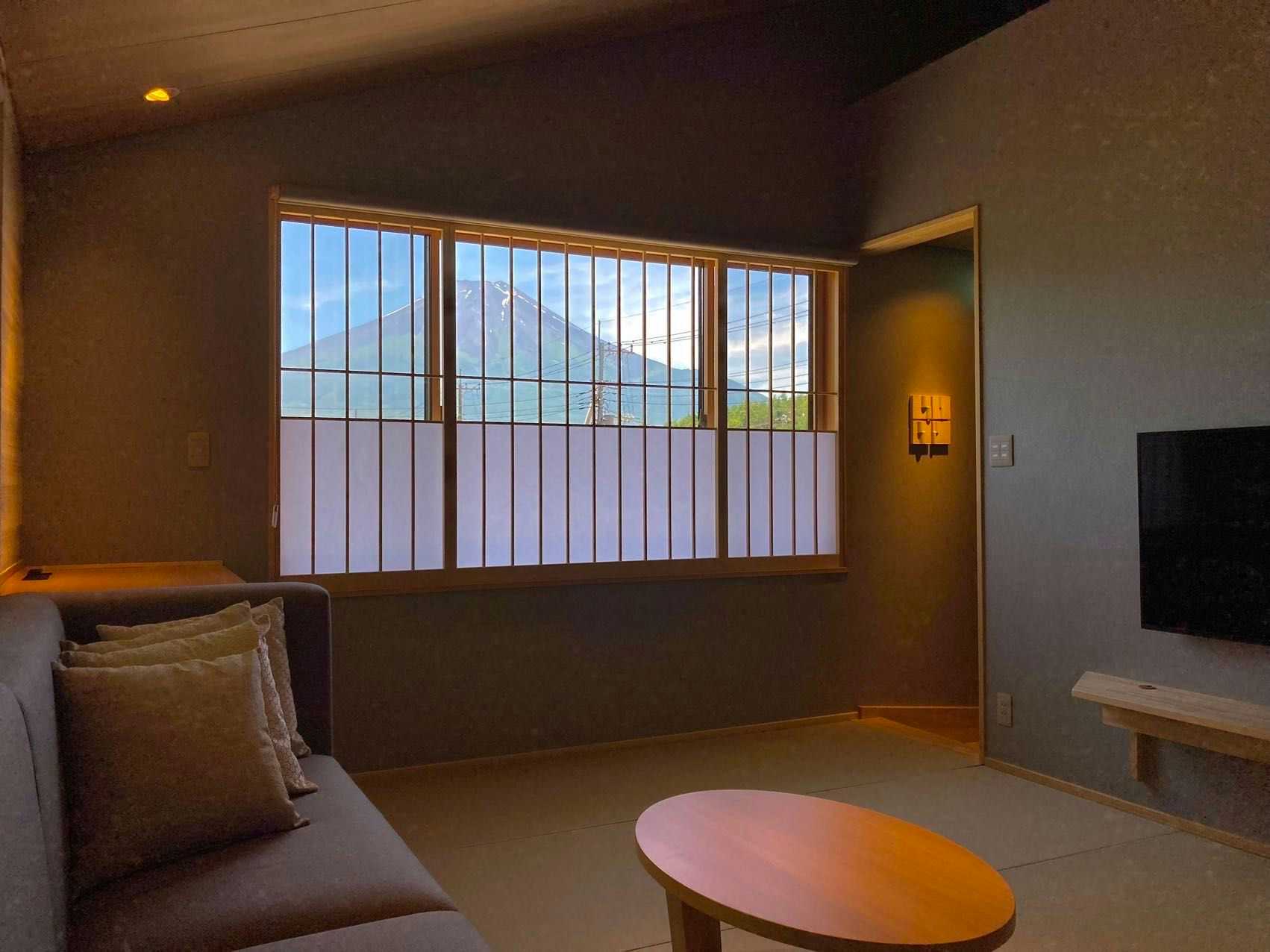 Hakkai konohanakan Sitting area with upholstered sofa, oval wooden table, wall-mounted screen, framed mountain view, and warm lighting