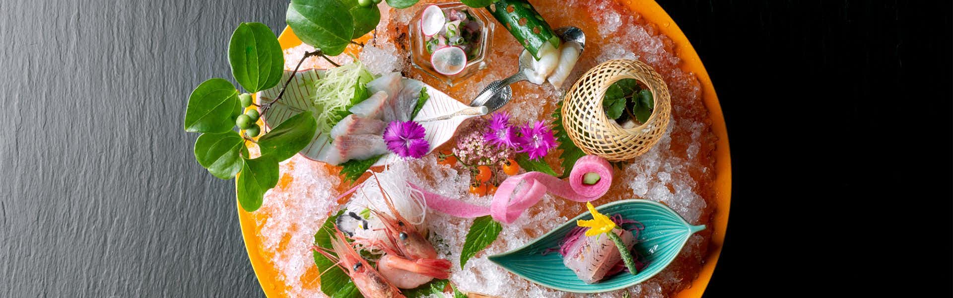 Noto Tsukumowan Hyakurakuso Food presentation on crushed ice with pink flowers, white and pink ingredients, fresh green herbs, woven basket, and teal serving dishes.