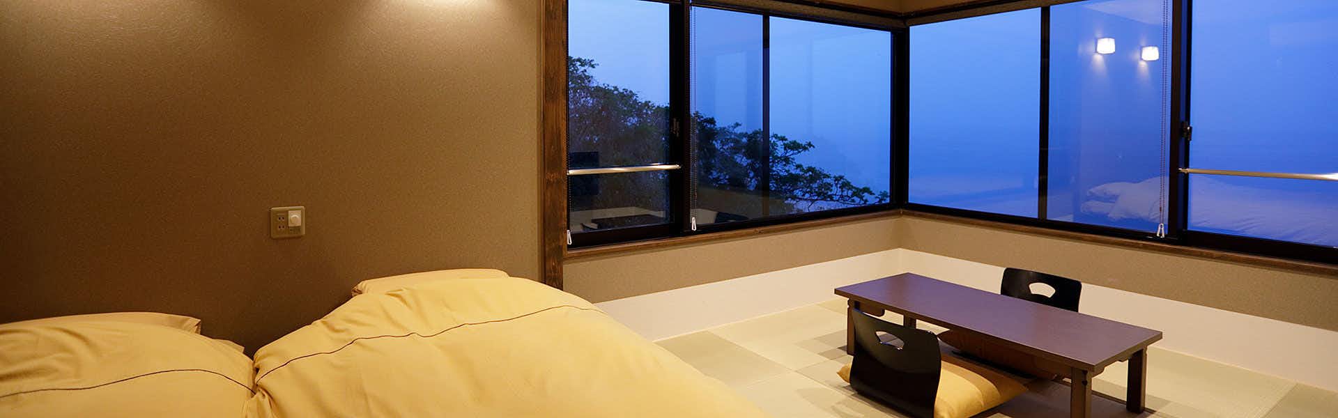 Hidden Hot Spring Inn Tsuki no Akari Tatami room with two futon beds, low table, floor chair, and corner windows overlooking trees and blue sea