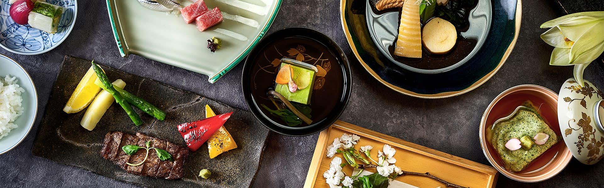 Yamatoya Besso Multiple ceramic bowls and plates with plated food, bread slices, herbs, and crystalline garnishes on dark surface