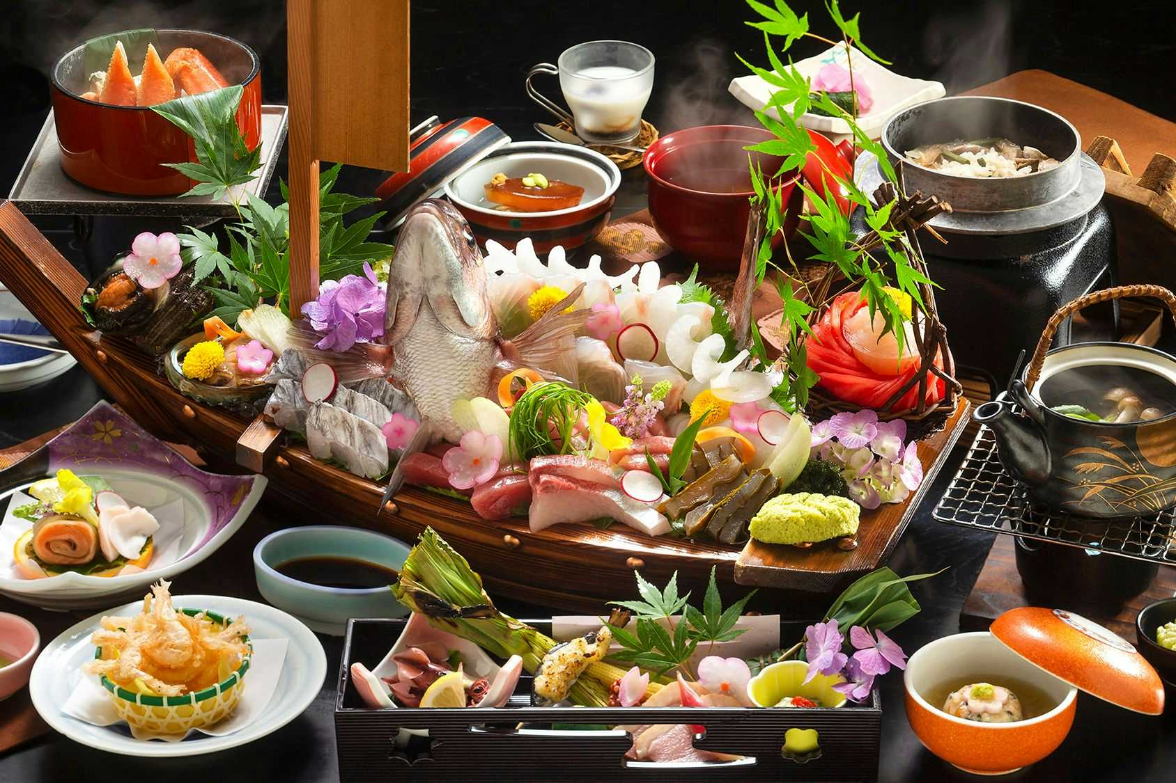 TOGEN Kurobe Unazuki Onsen Multiple bowls and plates with prepared items, flower garnishes in white and pink tones, beverages, and greens arranged on wooden trays.