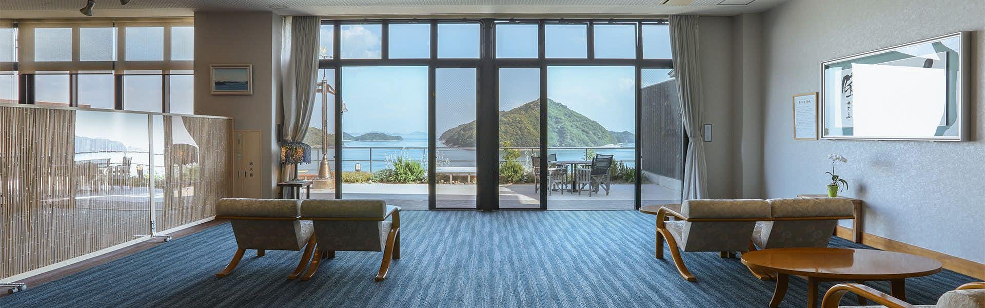 Kinoe Onsen Hotel Seifukan Lounge space with blue-striped flooring, wooden-frame chairs, dark-framed glass doors and windows overlooking water and distant mountains