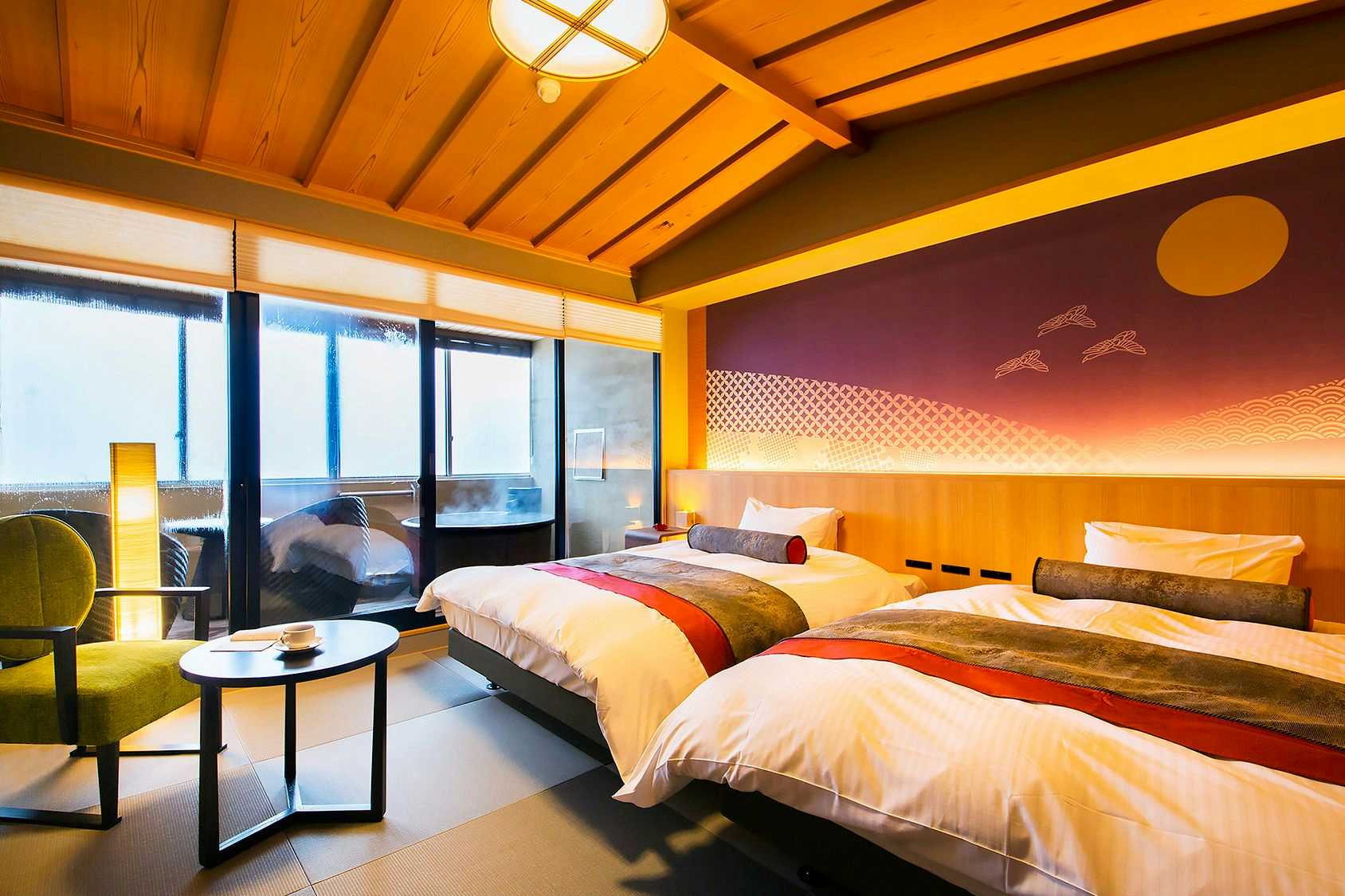 TOGEN Kurobe Unazuki Onsen Bedroom with wooden ceiling, large windows framing an orange-tinted sky, white bedding with red stripe accent, and round side table.