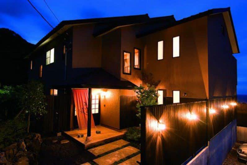 A Small Inn with Natural Hot Spring Flow: Ayu no Kaze Two-story exterior at night with glowing windows, dark brown walls, illuminated entrance, steps, and wooden privacy fence