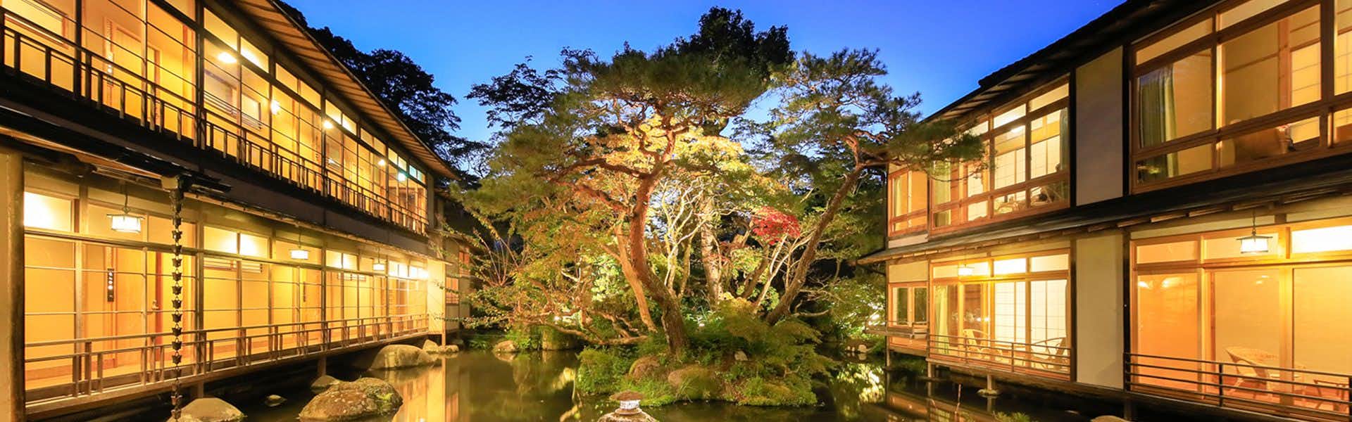 Arai Ryokan Large tree with thick trunk, wooden structures flanking on both sides, illuminated windows, water reflection at base, surrounding foliage and trees.