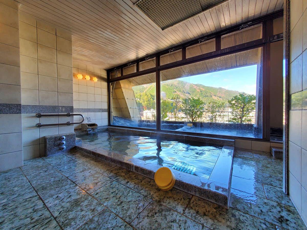 Gero Onsen - Hospitality and Flavor Accommodation, Kissenkan Chikusuitei Indoor stone bath with washing fixtures and a broad window framing wooded hills