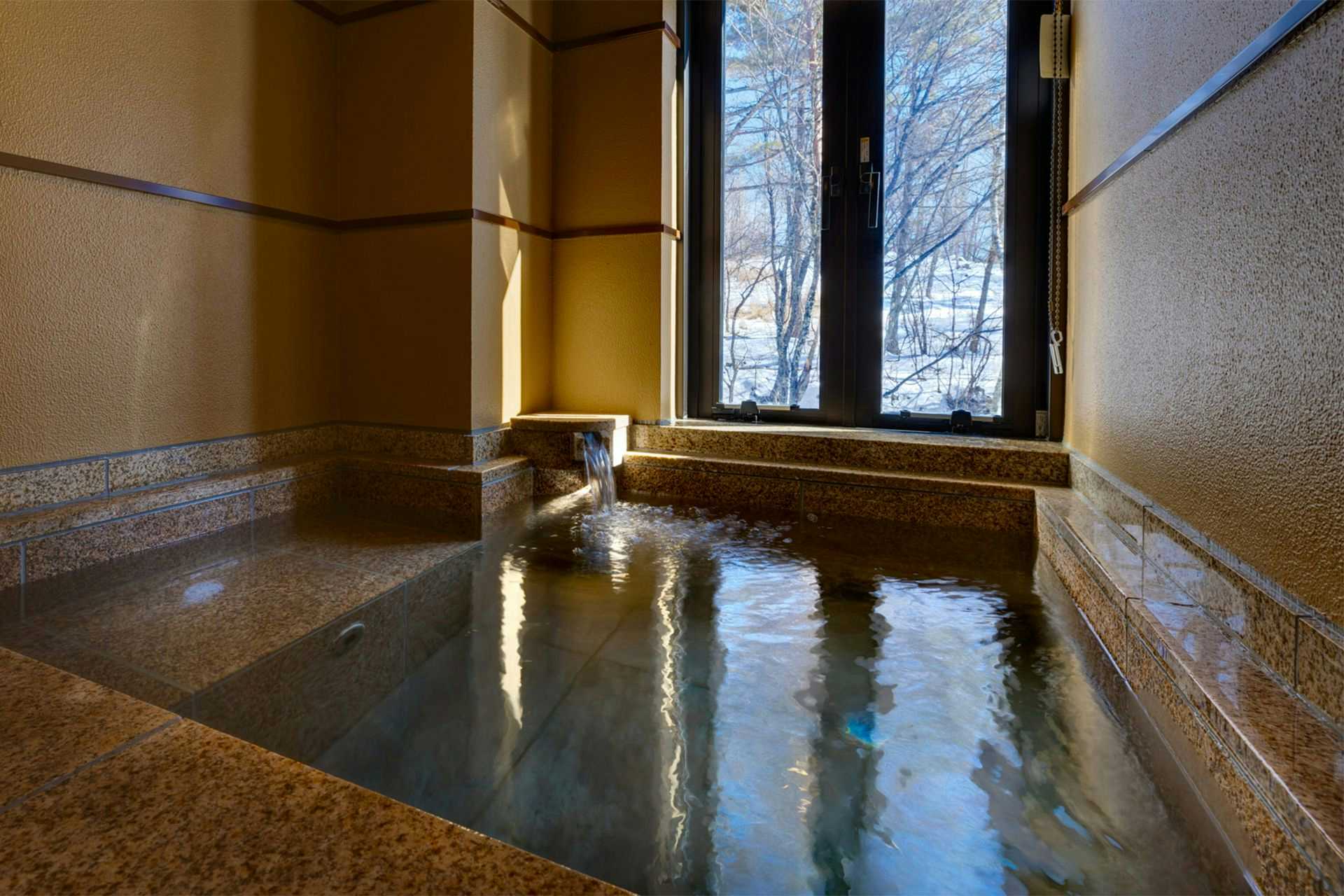 Bettei Tateshina Kaorukaze Indoor bathing pool with blue water and stone edges, beside floor-to-ceiling windows facing trees