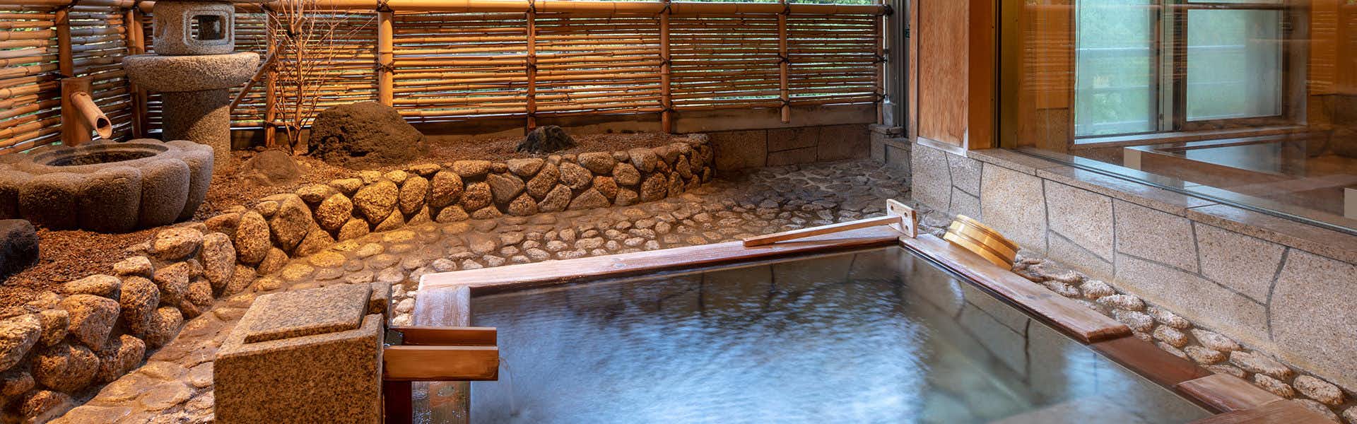 Bankokuya Rectangular bath with wooden trim beside stonework, bamboo screens, tiled walls, and a small stone lantern
