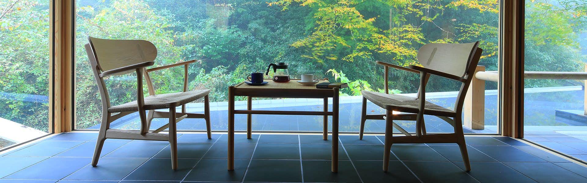 Okuyugawara YUI Wooden table and chairs with kettle and cups on surface, window framing forest landscape.