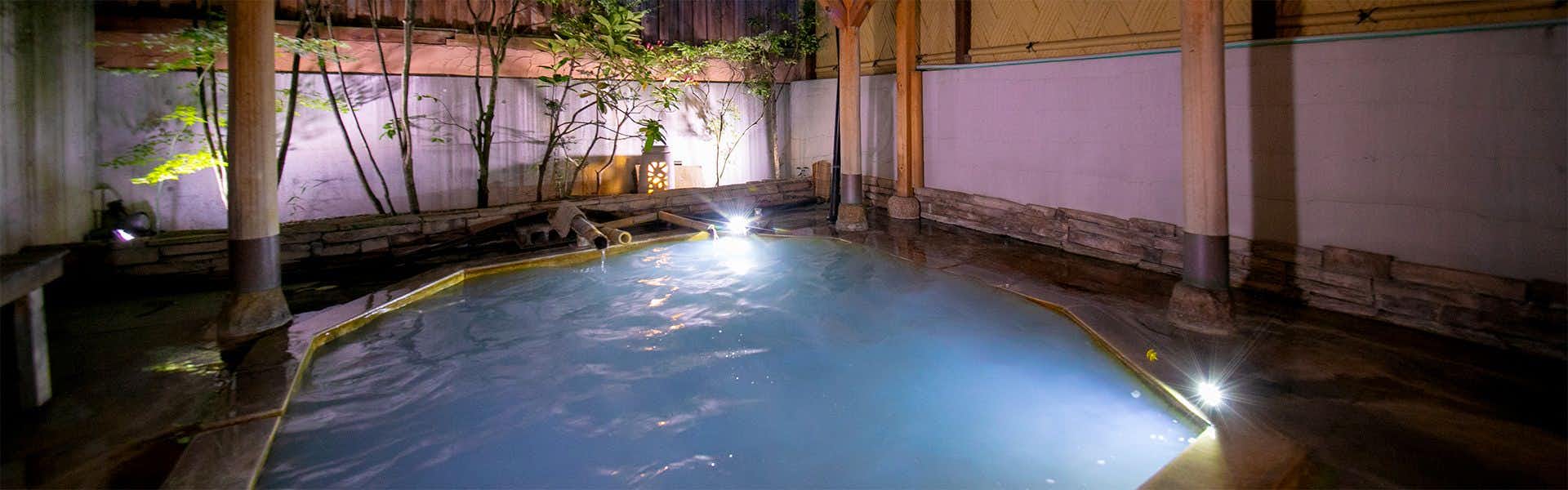 Yufuin Onsen Onyado Ichizen Large stone-lined open-air bath at night with wooden posts, bamboo water spouts, garden trees, and glowing underwater lights