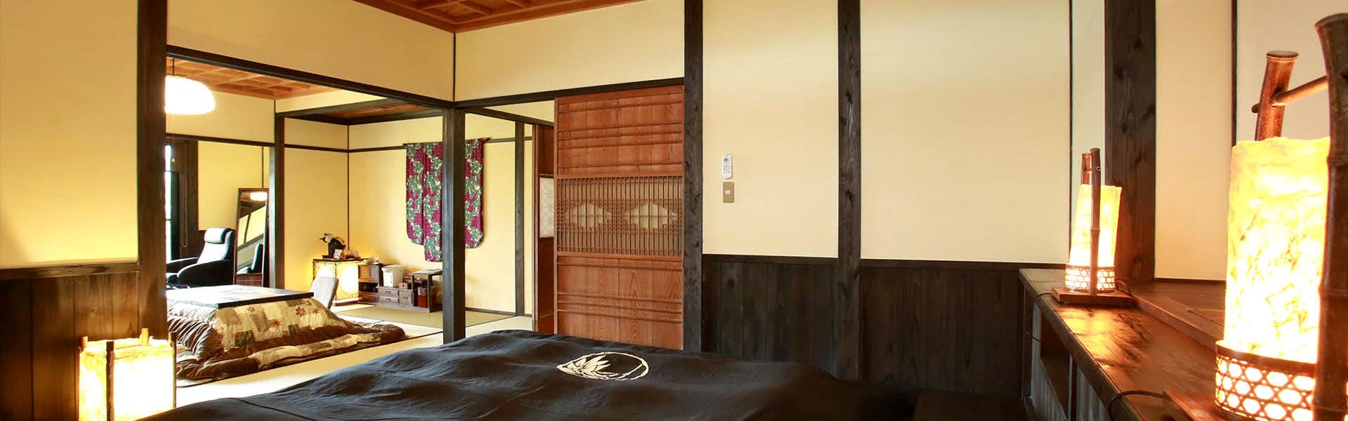 Minami Aso Onsen Iyashi no Sato Hatago Seiwa Traditional guest room with dark-covered bed, adjoining tatami sitting area, low covered table, timber trim, and warm lanterns