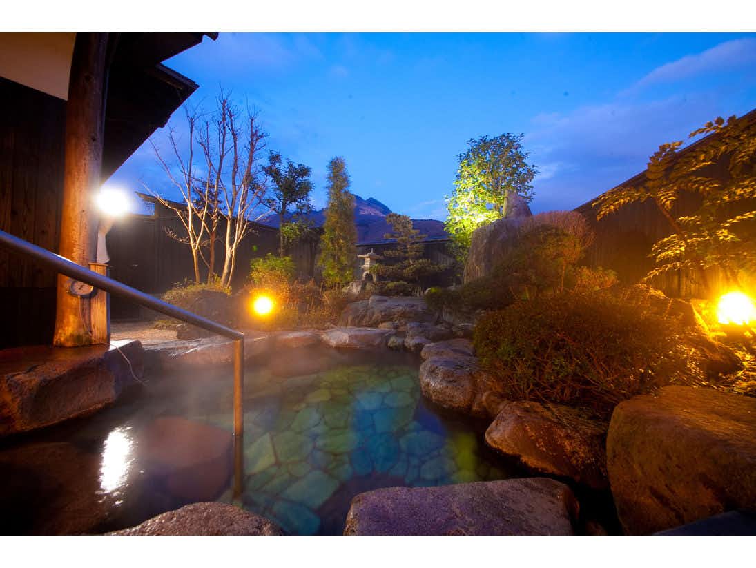 Yufuin Yawaragi no Sato Yadoya Outdoor bath at night with warm lighting, natural rock formations, illuminated trees, and steaming water surface.