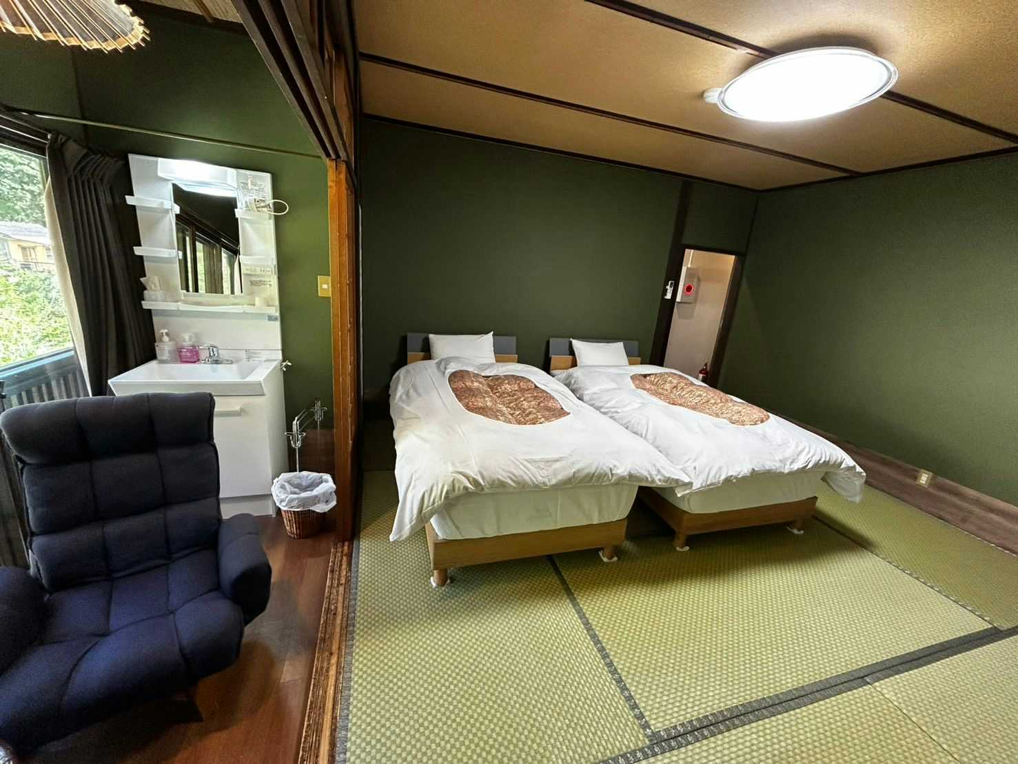Ryokan Hakuunsou Bedroom with white bedded frame, patterned pillows on tatami flooring, dark green accent wall, and black upholstered chair.