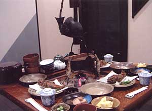 A meal arranged around a sunken hearth beneath a carved wooden fish hanger.