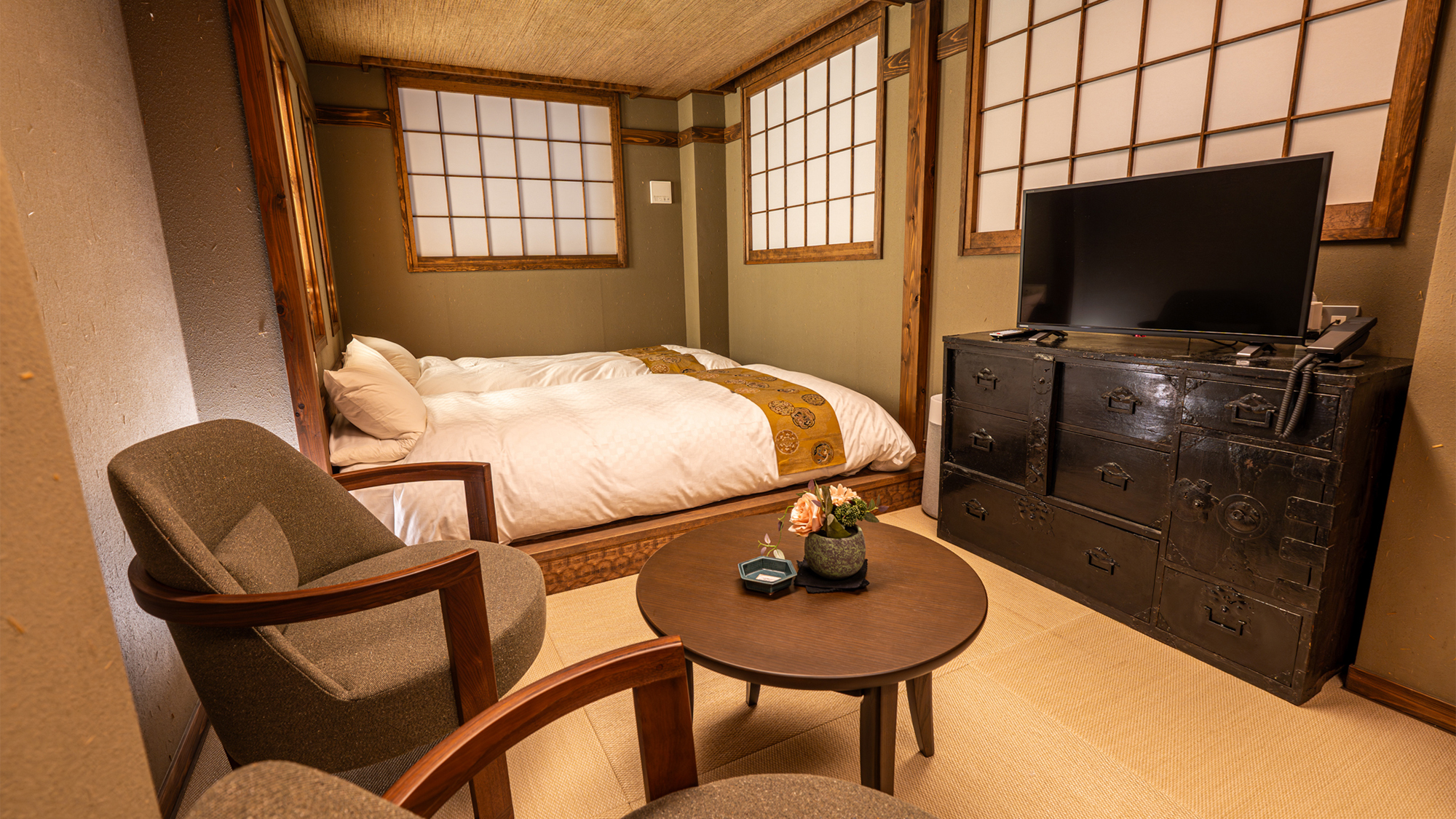 A traditional style room has two futons, shoji screens, two armchairs, a round table with flowers, and a television on a dark cabinet.