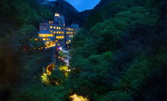 Illuminated multi-storey hotel across a densely wooded valley at dusk.