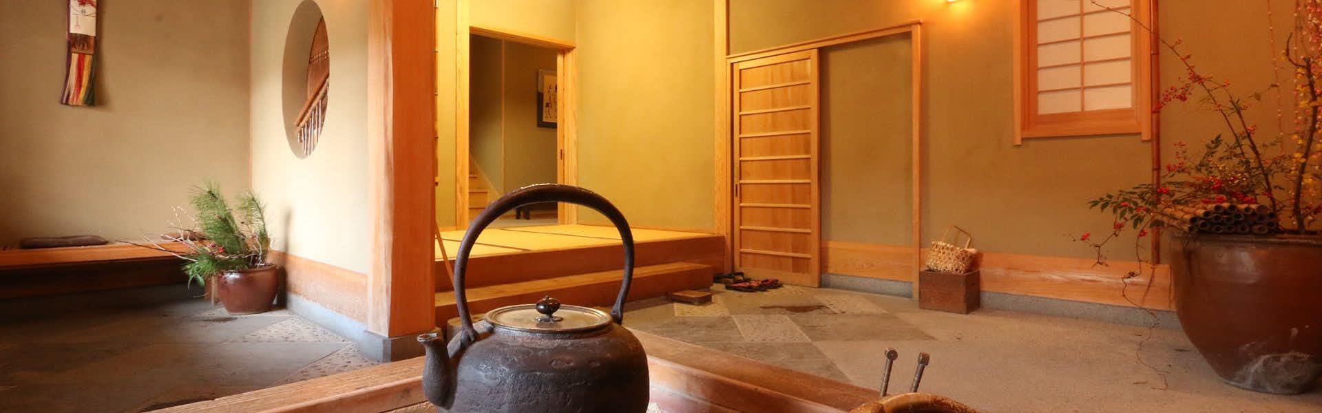 Marucho Ryokan Wooden interior with black metal kettle, wooden panel doors in warm tones, potted plant, wooden floor, and sunlight.