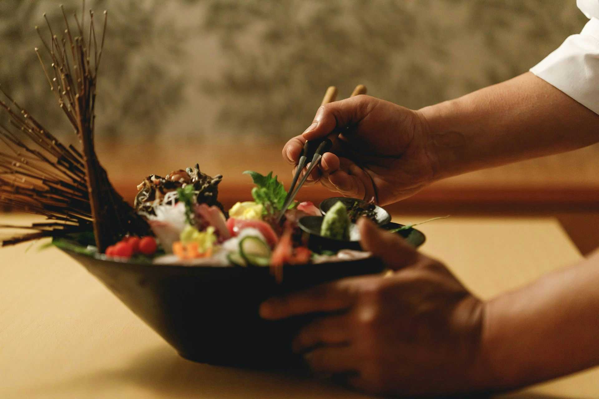 Lakeside Inn Morimoto Hand with chopsticks over black dish containing prepared items, greens, and colorful elements, warm sunlit presentation.