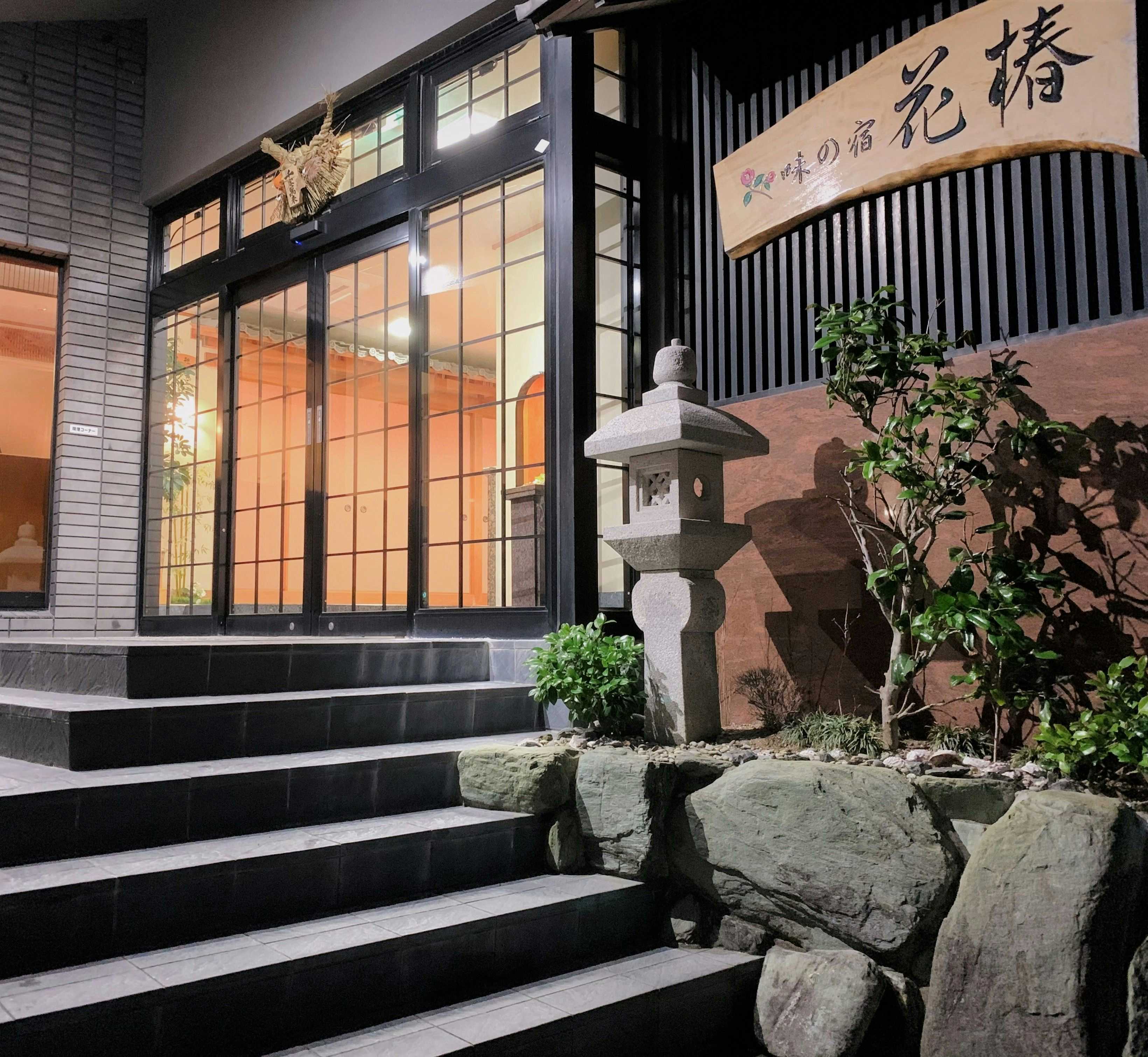 Aji no Yado Hanatsubaki Entrance with dark wooden door frame with vertical slats, stone steps, decorative stone lantern sculpture, and potted plants.