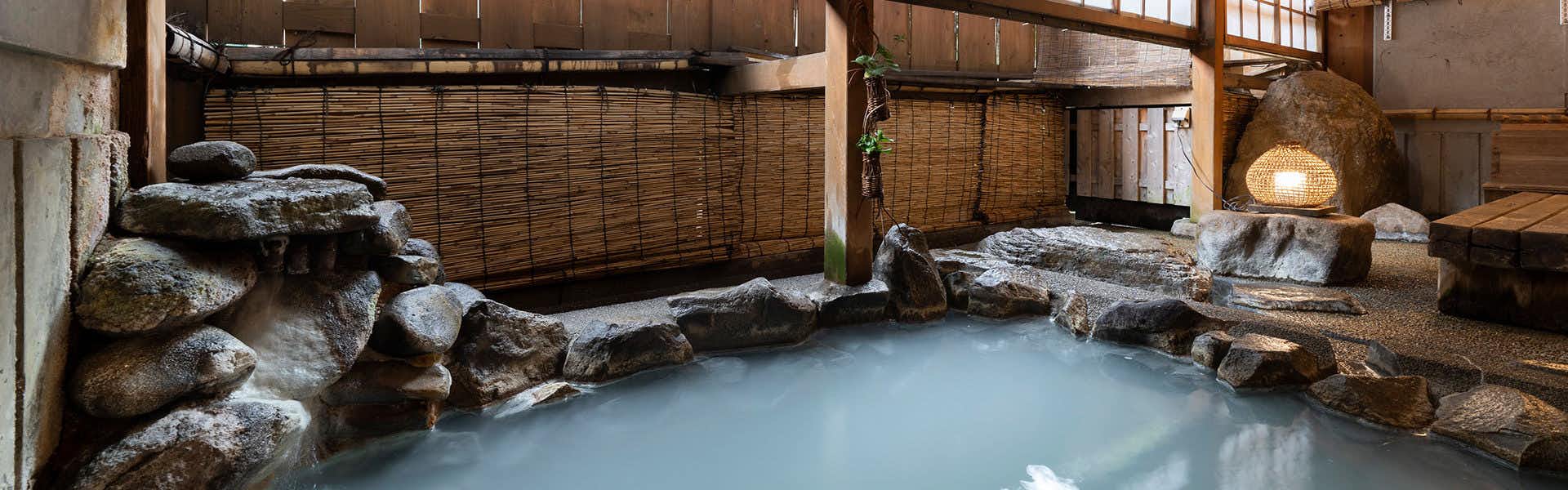 Nozawa Onsen Kawahichi-ya Ryokan Indoor bath with large natural rocks, clear water, wooden railings, and stone walls enclosing the basin.