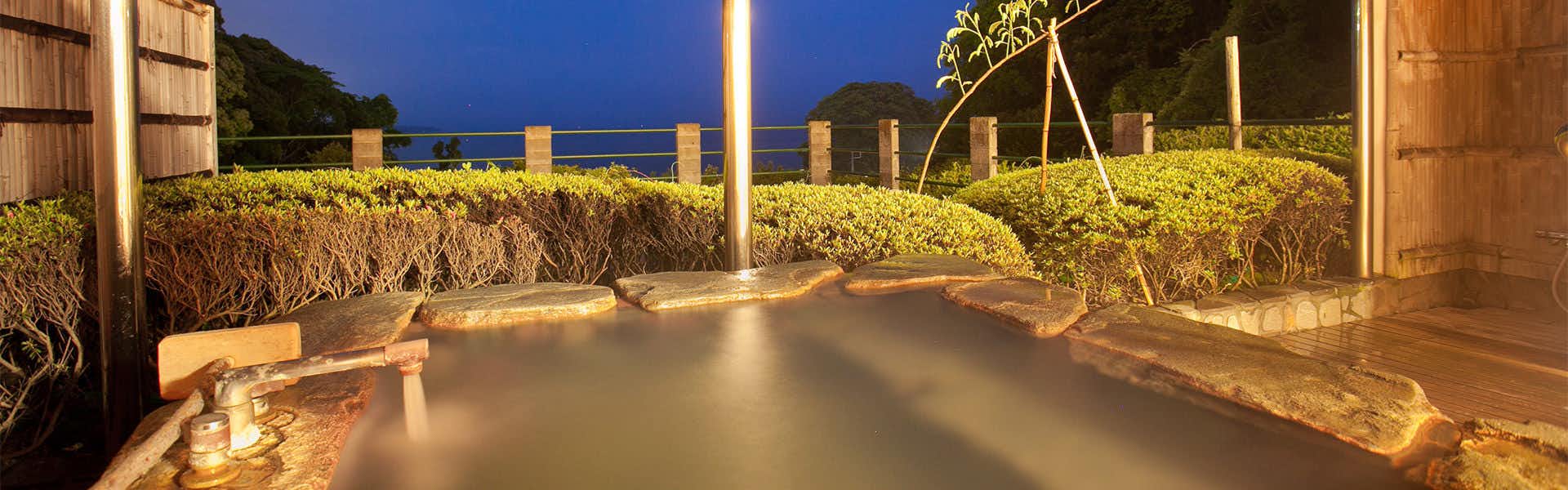 Isaribi Inn with All Rooms Featuring Open-Air Hot Spring Baths Outdoor bath with wooden pillars and landscaped plantings, ocean view visible in evening light.