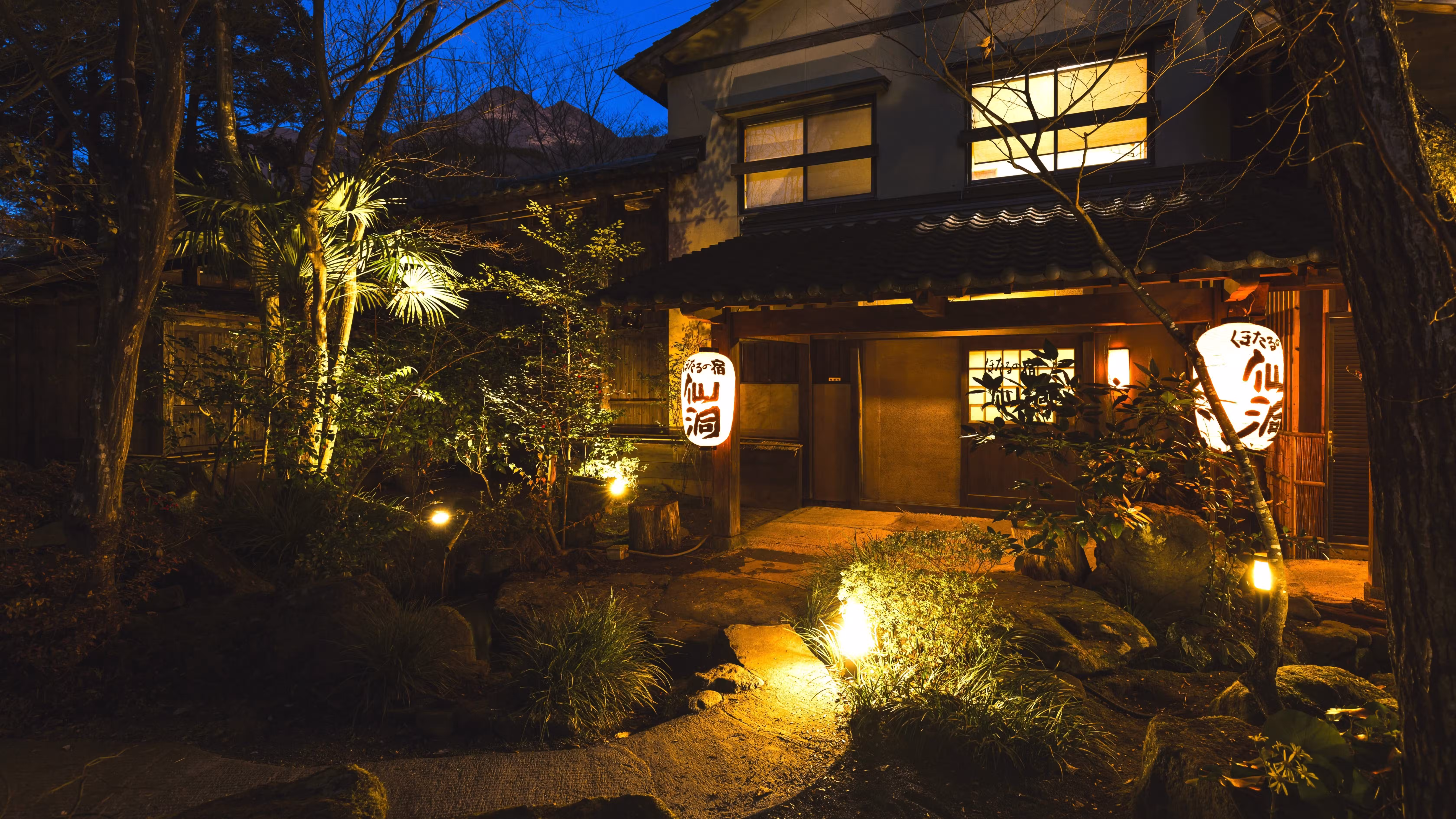 Hotaru no Yado Sendou with this scene, where the ryokan’s timber entrance glows along a stone garden path after dark.