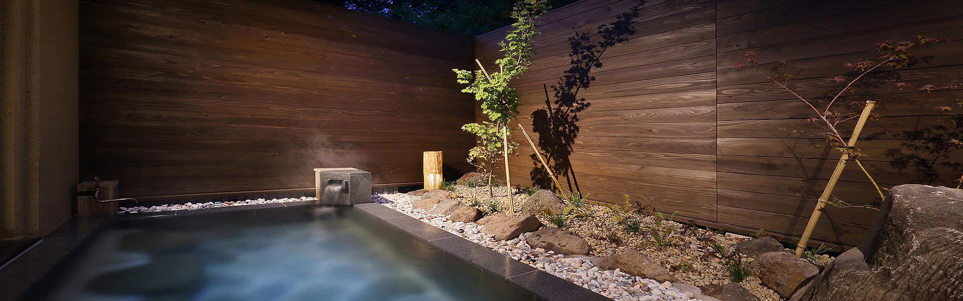 Oyado Mori no Ne Rectangular outdoor bath glowing at night beside white gravel, small trees, rocks, and dark horizontal wood walls.