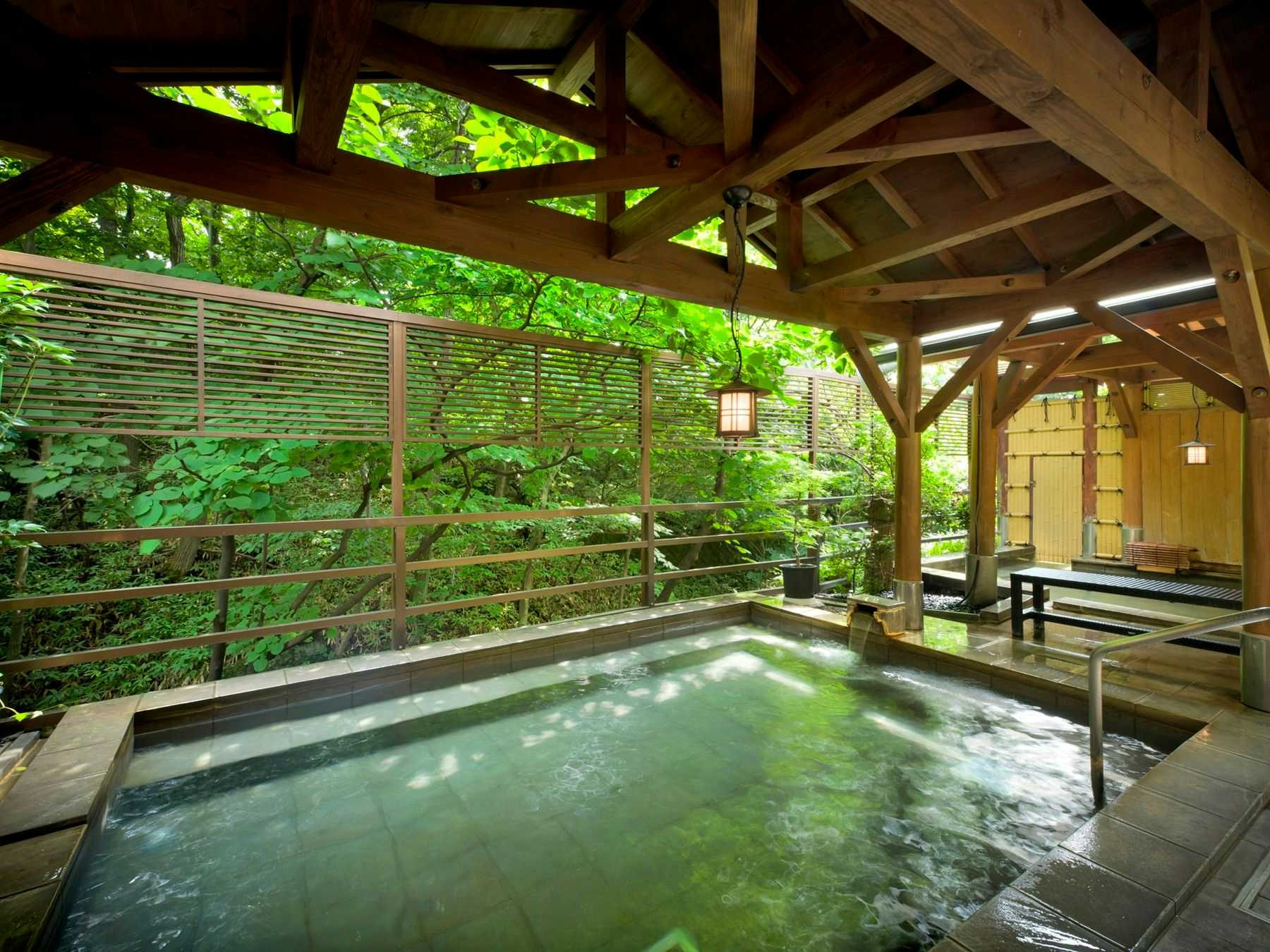 Kokoro no Oyado Jizai-so Roofed rectangular bath with dark decking, metal handrails, timber beams, and dense green forest beyond