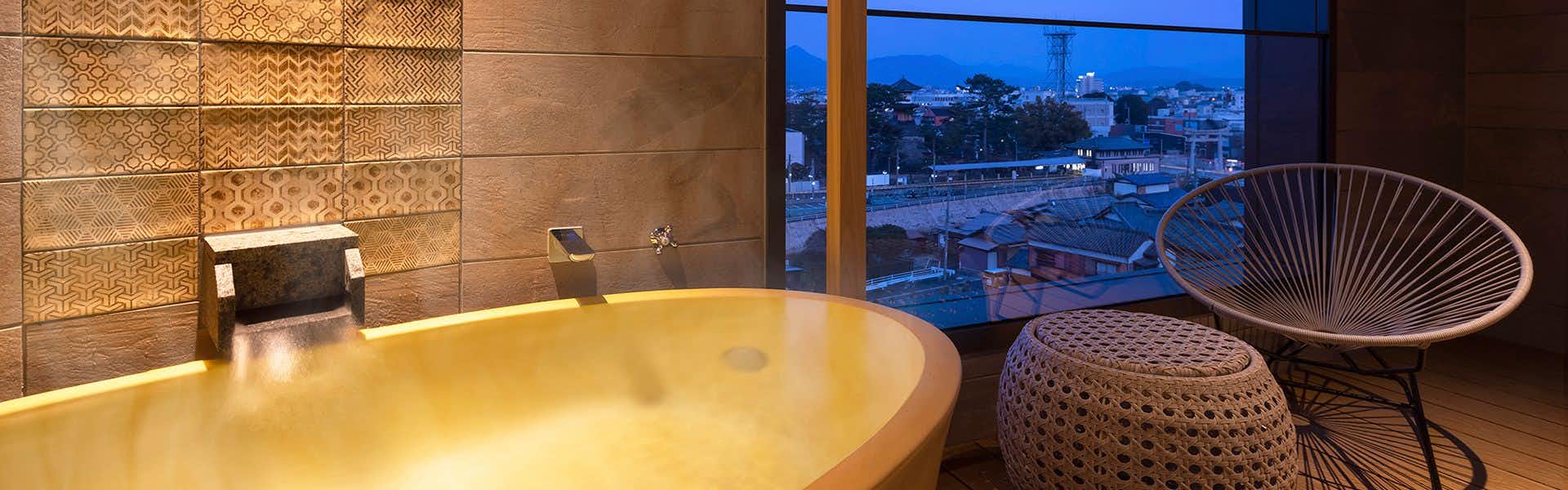 Yumoto Konpira Onsen Hananoyu Koubaitei Round golden bathtub with city view through expansive window, dark woven chair, and wooden wall panels.