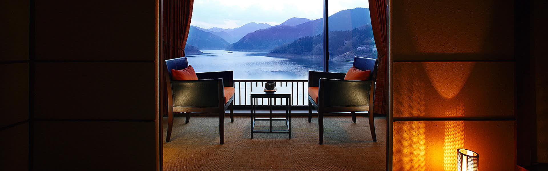 Akane Saian Sansui Two red-orange chairs and small table positioned before tall windows overlooking mountains and a reflective lake.