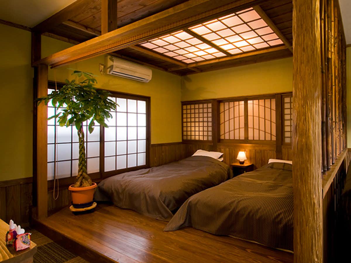 Kaede no Shoja Bedroom interior with dark wood bed, brown bedding, geometric wooden ceiling skylight, mustard yellow walls, potted plant.