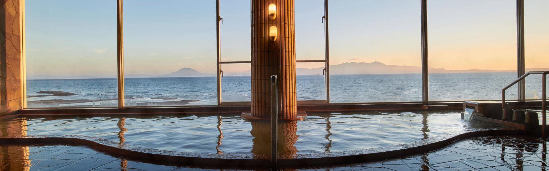 Koran no Yu Kinkouro Indoor bath with blue-tinted water, wooden support column, glass enclosure, windows showing water view.
