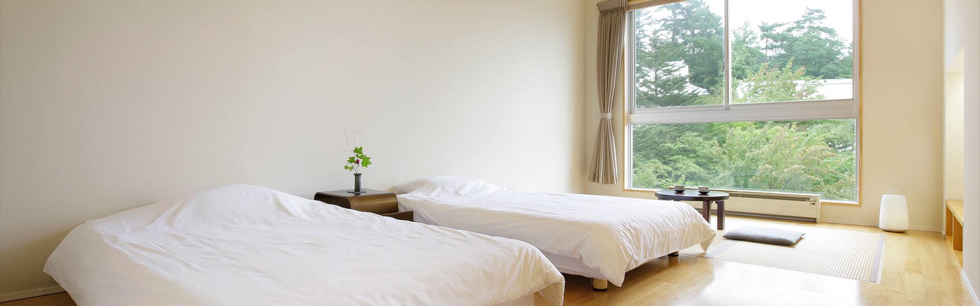 Kusatsu Hotel Annex Wata no Yu Bedroom with white bedding, large windows with beige curtains overlooking trees, wooden furniture, and potted plant.
