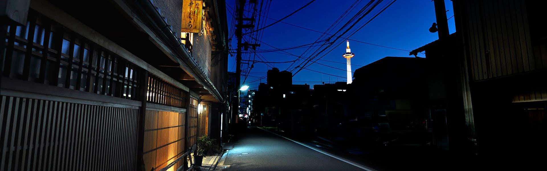 Izuyasu Traditional street at dusk with dark wooden facades, glowing lanterns, overhead wires, and a distant illuminated tower