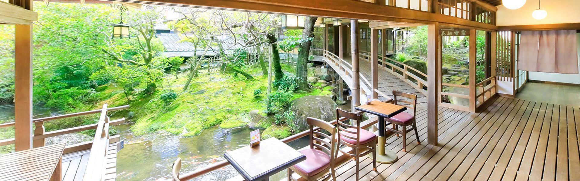Arai Ryokan Interior with wooden beams, wooden flooring on platform, cushioned seating on low furniture, wooden posts, large windows overlooking garden with moss and green vegetation, stream visible.