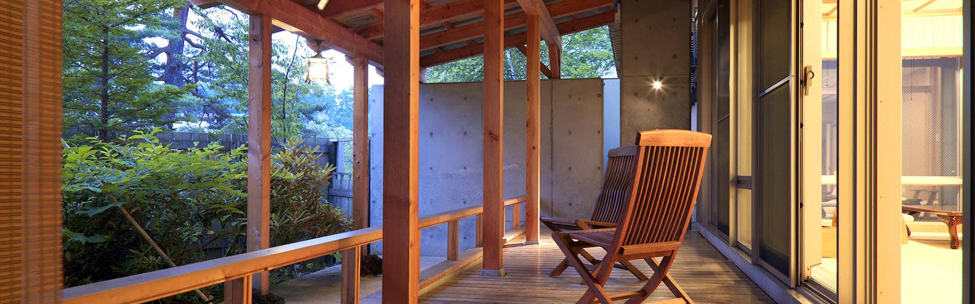 Kusatsu Onsen Bouun Wooden deck with large glass doors, wooden railings and chair, forest view illuminated by warm evening light.