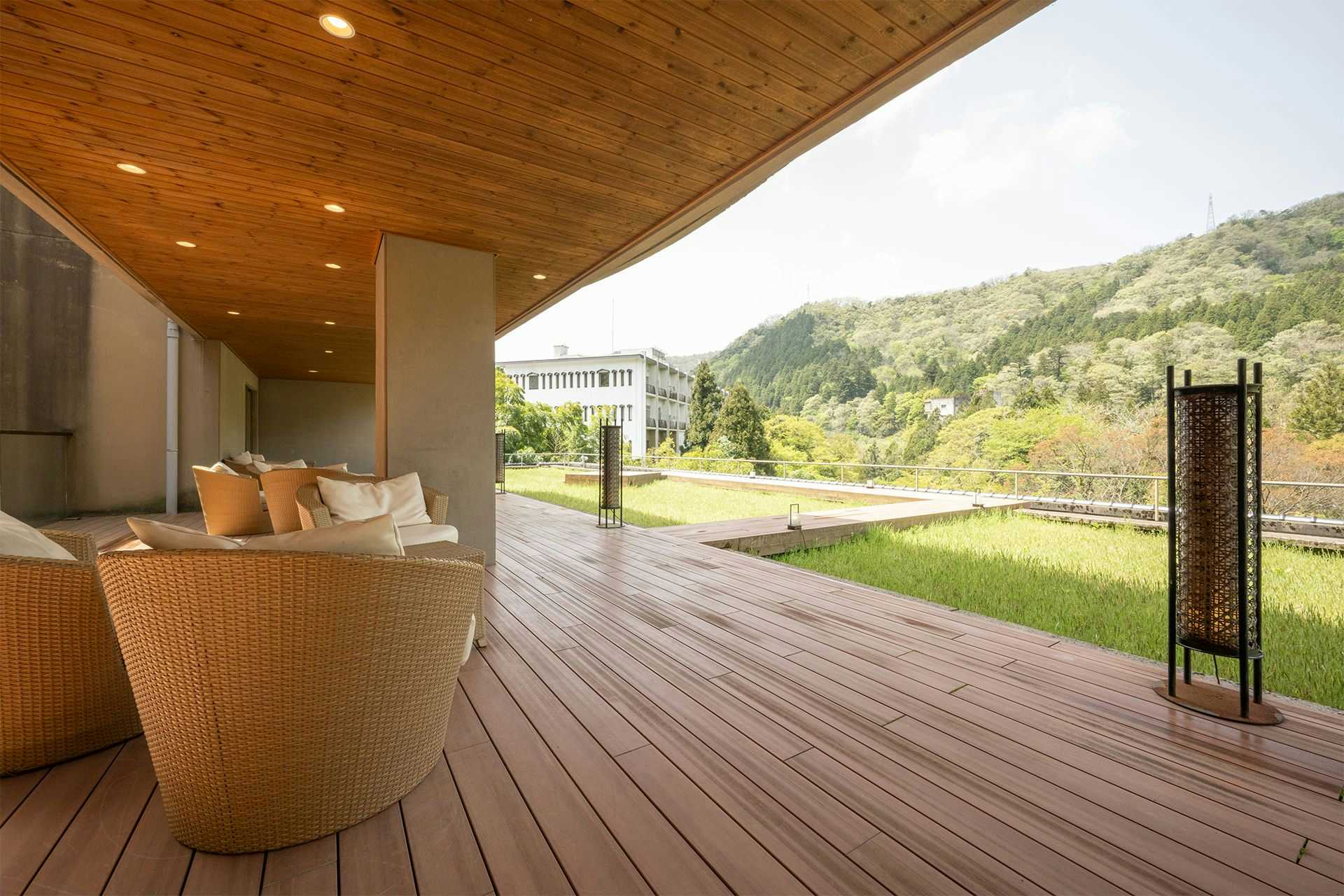 Kuriya Yasohachi Covered terrace with wicker chairs, wood decking, deep eaves, tall lanterns, grass, and a green hillside